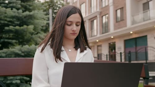 Woman remotely working sitting on bench at city street using laptop.