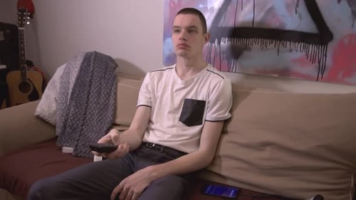 A Young Man Holding A Remote Control And Pressing The Button While Sitting On The Sofa At Home. - cl
