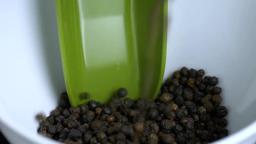 Close-up of whole black peppercorns sliding in a white bowl. The bowl fills up slowly, a green plast