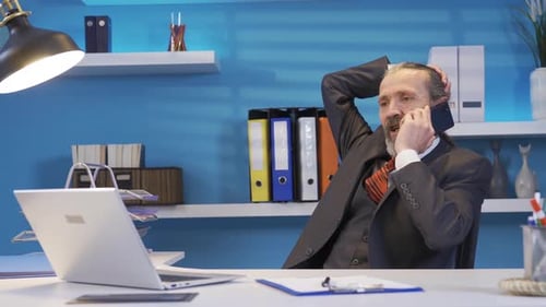Senior mature businessman sits at his desk in the office and talks on the phone.
