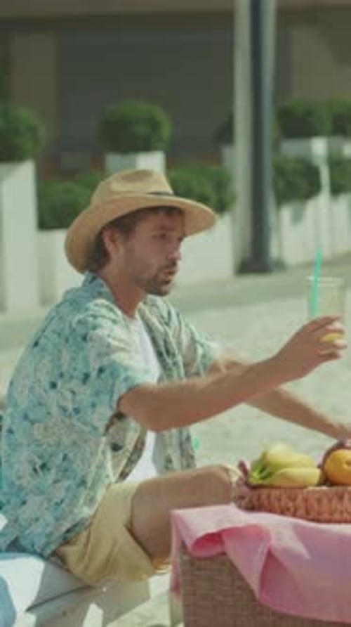 Enjoying Fruit at Sunny Beach Male Individual Raises Cup with Fruit Basket Behind Beneath Bright