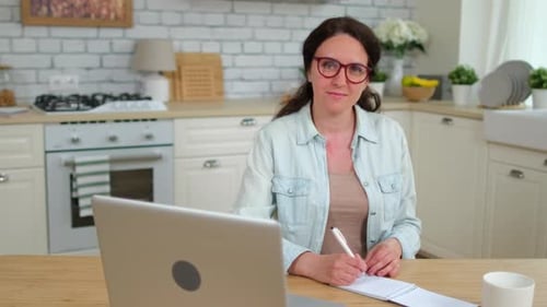 Woman Taking Notes at Kitchen Table with Laptop