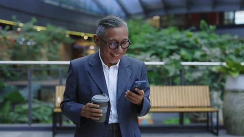 Cheerful Senior Man Goes to the Office with Coffee and Watches Funny Videos on the Phone