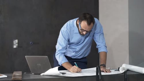 Man Working on Blueprints with Laptop Indoors