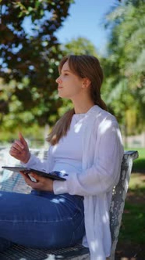 Young Female Interacting with Digital Tablet While Resting on Wooden Park Bench Displaying Animated