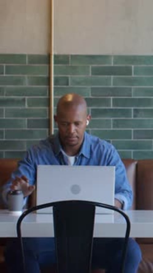 Vertical video: Looking at laptop, man in blue shirt typing and sipping coffee cup for work at cafe