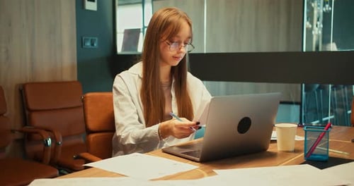 Focused Blonde Girl in a Business Suit Takes Notes and Sits in Front of a Laptop with a Table in the
