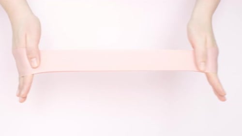 Faixa elástica de fitness esticando mãos femininas em close-up de fundo rosa.