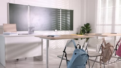 Green chalkboard, desks and chairs with backpacks in classroom. Camera moving right