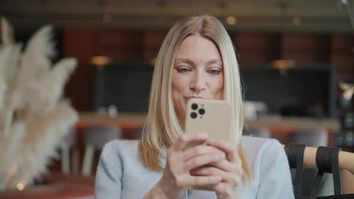 Portrait of Attractive Young Blond Woman Holding and Using Mobile Phone Sitting in Cafe Texting on