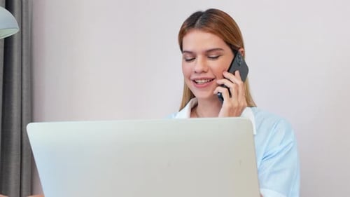 Young caucasian businesswoman talking with smartphone and working on laptop computer in home office.