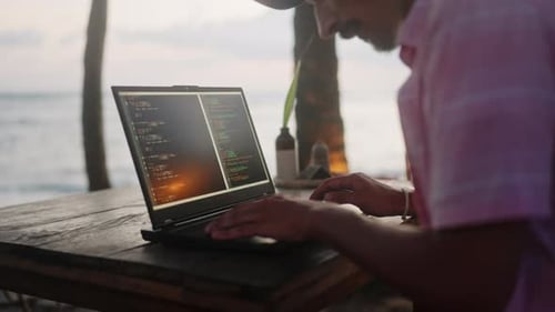 Male Developer Types Code on Laptop at Beachside Cafe Sunset Behind with Ocean View Pro Works Remote