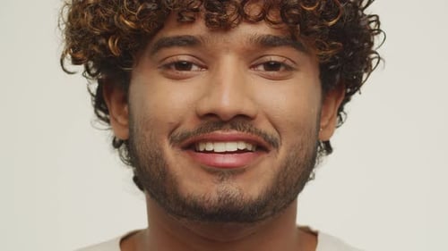 Hübscher junger Mann fängt an zu lachen und schließt die Augen. Lockiges gemischtes männliches Model posiert auf weißem Hintergrund