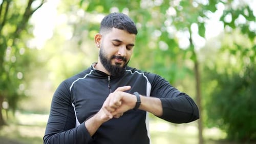 A bearded male runner stands in an urban city park and looking smart watch. Handsome athlete using