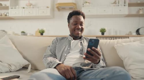 Carefree Black Man Sitting on Sofa in Living Room Using Smartphone Pastime Portrait of Positive