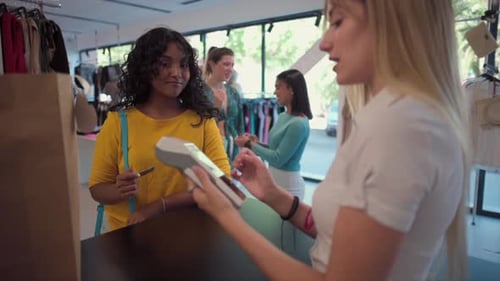 young woman in her 20s makes a contactless card payment at a clothing store counter while the cashie