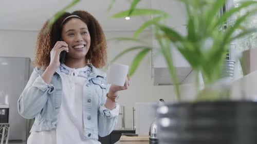 Happy biracial woman talking on smartphone, laughing and holding coffee in kitchen, slow motion