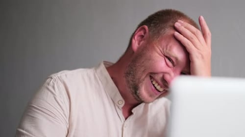 Adult man in a good mood laughs looking at the monitor screen against a light wall, close up.