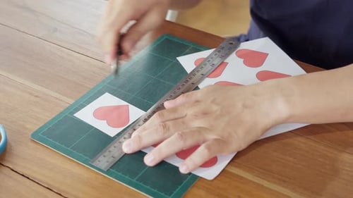 Close-up of hands cutting heart shape paper using utility knife cutter with ruler on cutting mat.