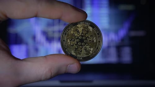 Hand Holding Cardano Coin Against Laptop Screen