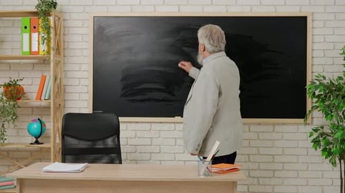 Aged Man College Professor Standing at Desk in Classroom in Front of Chalkboard Writing Chemistry