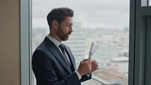 Businessman Folds Paper Airplane by Office Window Contemplatively