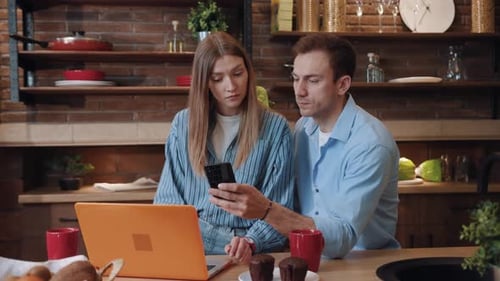 Couple Using Laptop and Smartphone at Kitchen Table