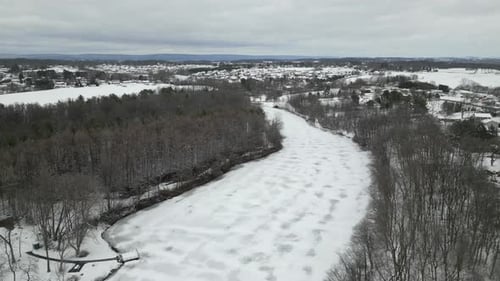 A cinematic high-angle drone shot capturing a wide frozen river surrounded by snow-covered trees