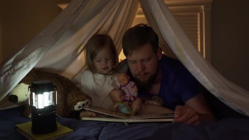 Father reading to toddler daughter at bedtime in blanket fort