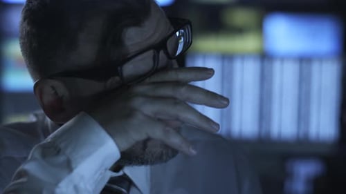 Manager is working on a computer in the evening in a dark office room filled with display screens.