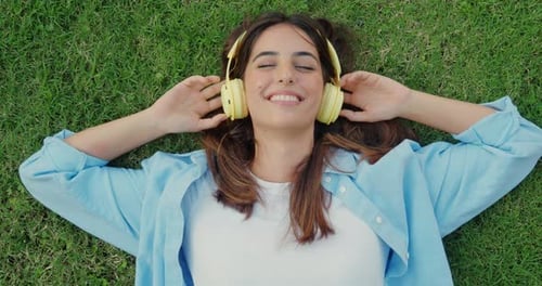 Junge fröhliche Millennial-Frau liegt auf dem Gras und hört Musik mit Kopfhörern