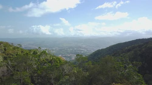Drone flies beyond sunlit forest mountain peak, to reveal city
