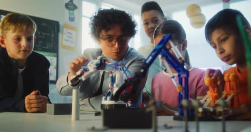 Team of Talented Kids Sitting at Table Studying Robotic Arm Model with Teacher
