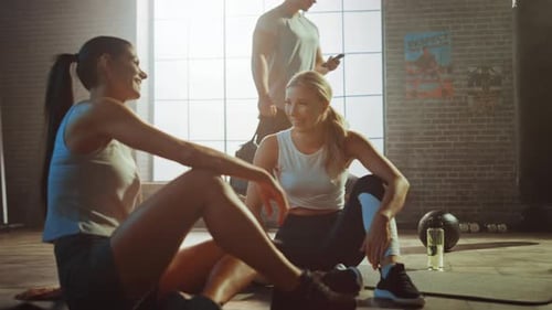 Two Beautiful Fit Athletic Girls Sit on a Floor of Industrial Loft Gym. They're Happy with their Tr