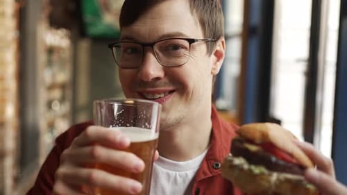 Bearded Man Wearing Glasses Sipping Craft Beer Eating Burger with Relaxed Smile at Wooden Restaurant
