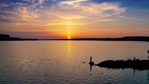 Silhouette of a fisherman with fishing rod standing on stone near the river at sunset.