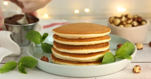 Woman spreading chocolate paste onto pancake at light wooden table, closeup