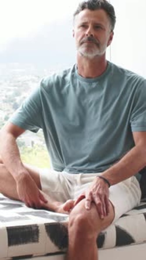 Vertical video: Reacting to window light, mature man leaning on bench at home, teal tee other foot