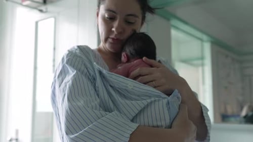 Mother tenderly kissing newborn baby’s head, wrapped in a striped hospital gown, capturing the