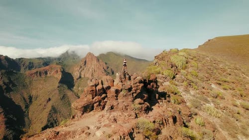 A woman stands atop a rock amidst rugged mountains. Distant peaks, mostly shrouded in clouds, sugges