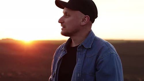 Side view of male farmer inspecting cultivated plowed field at sunset and holds digital tablet