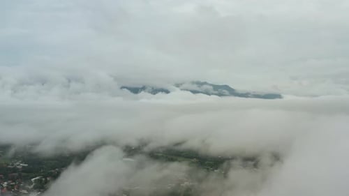 Aerial Flying above Clouds with town beneath. Mountain Peak sticking up above clouds in Distance.