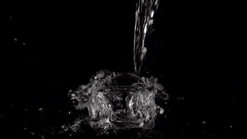 Ein Wasserstrahl fällt in einen Glasbehälter auf schwarzem Hintergrund und erzeugt einen Spritzer Wasser
