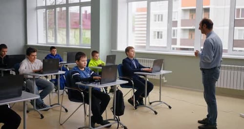 Jeunes élèves assis à des tables avec des ordinateurs portables dans une salle de classe assistée par ordinateur, professeur parlant