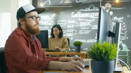 In the Creative Agency Videographer in a Cap Works on Personal Computer with Two Monitors, He's Doi
