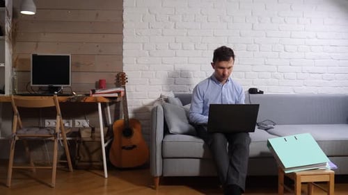Man Working From a Grey Sofa with a Laptop in a Modern Living Room Focused on Tasks and Illustrating