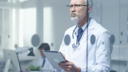 Senior Doctor Thinking About Patient's Diagnosis and Typing on a Tablet Computer. His Office is Bri