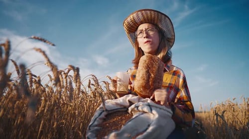Young Girl Farmer in Sun Hat Eats Loaf of Bread Biting It Drinking Milk Sits on Wheat Field with