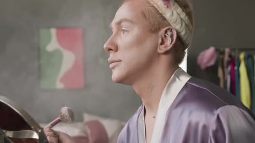 Hombre queer soplando el polvo de la brocha mientras se maquilla en casa