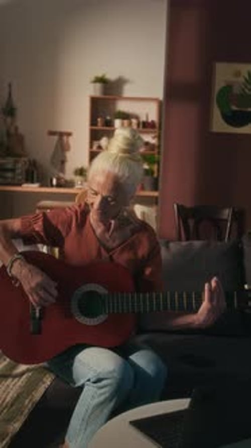 Mujer mayor que aprende a tocar la guitarra sentada en el sofá frente a una computadora portátil abierta